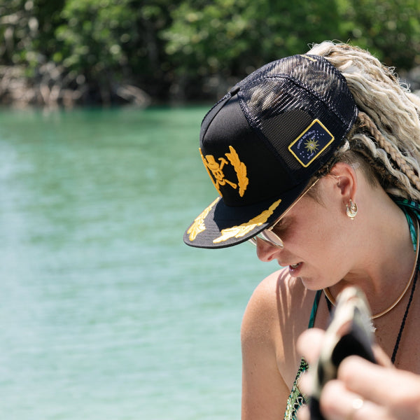 A Pirate's Life for Me Hat in Black — lifestyle shot on model on the water, yellow pirate skull emblem front, Conch Republic patch on side | Lost Boy Creations Key West