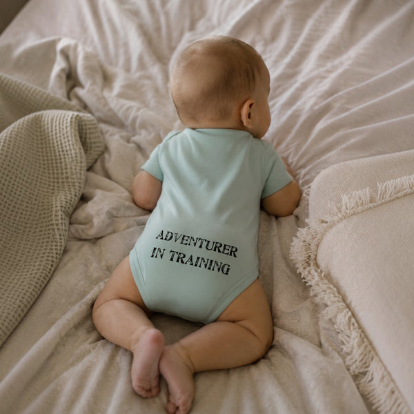 Kid's Lost Boy in Training Onesie in Fern — lifestyle shot, baby wearing onesie showing "Adventurer in Training" back print, laying on soft bedding | Lost Boy Creations Key West