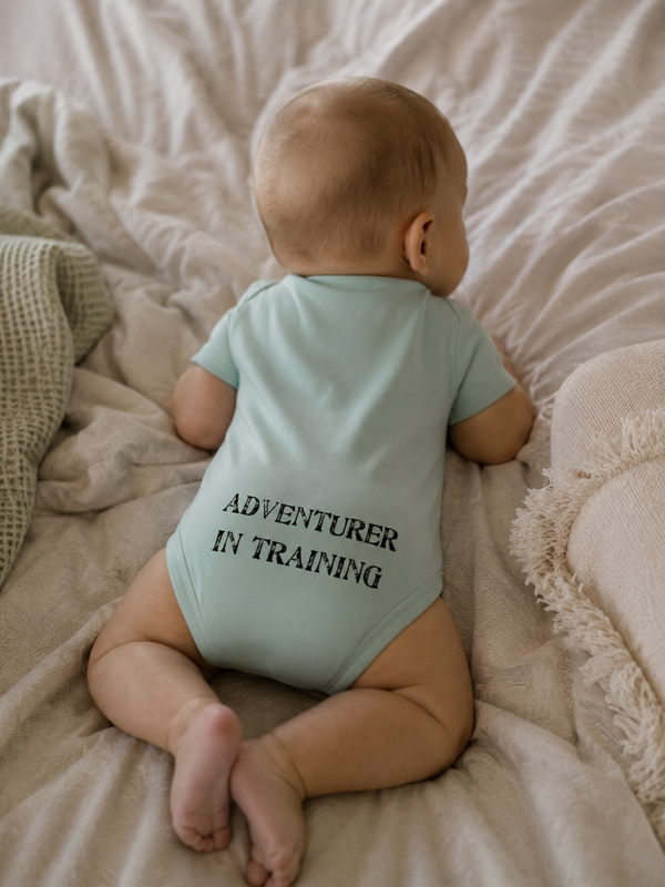 Kid's Lost Boy in Training Onesie in Fern — lifestyle shot, baby wearing onesie showing "Adventurer in Training" back print, laying on soft bedding | Lost Boy Creations Key West