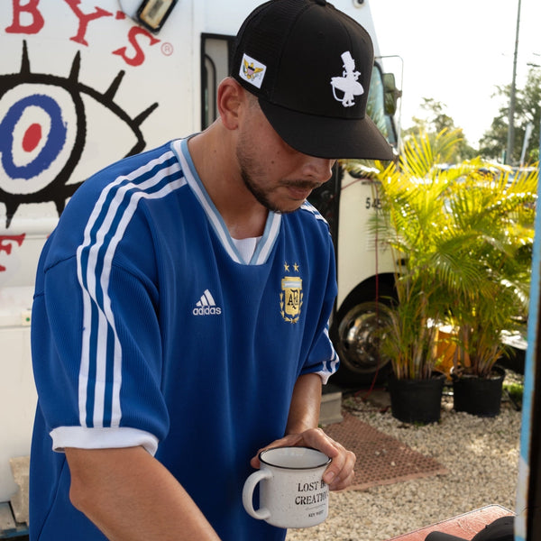 Flex'd all black low crown stretch-fit hat worn by a man in a blue Adidas shirt holding a coffee cup near DJ equipment outdoors | Lost Boy Creations Key West