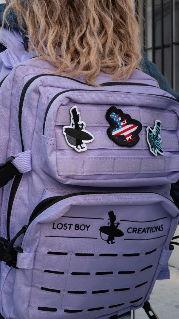 Girl wearing Lost Boy Creations 25L Lavender Black adventure backpack with black silhouette American flag and wave runner patches on front velcro panel | Lost Boy Creations Key West
