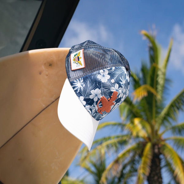 Lei'd Back navy floral low crown trucker hat displayed on a surfboard with palm trees in the background | Lost Boy Creations Key West