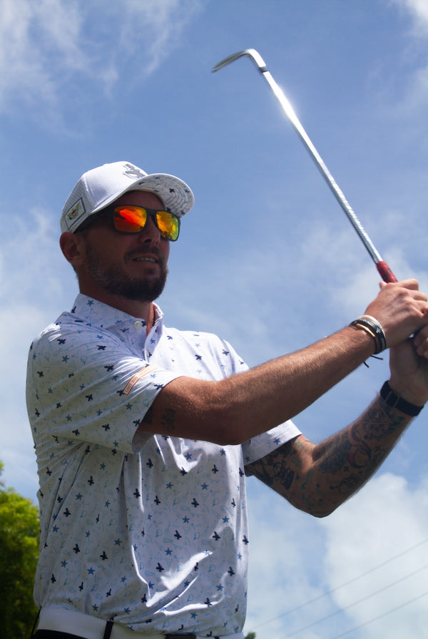 LBC Golf Polo white lifestyle shot worn by man mid golf swing outdoors with all-over surfer silhouette and star pattern and LBC trucker hat | Lost Boy Creations Key West