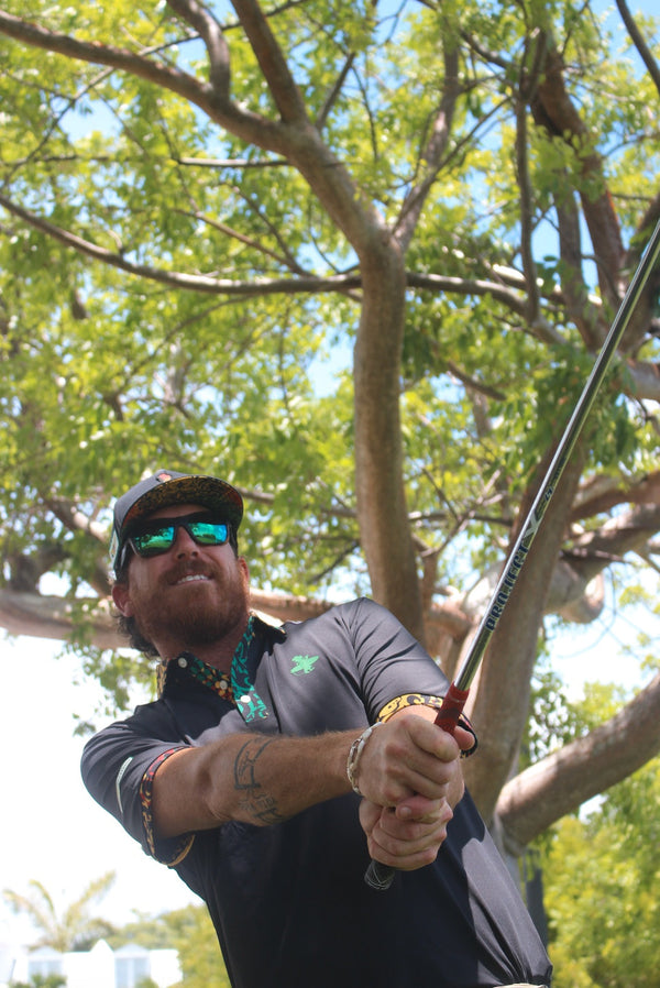 Rasta Golf Polo black lifestyle shot worn by man mid golf swing outdoors with green LBC logo and Rasta-print collar visible | Lost Boy Creations Key West