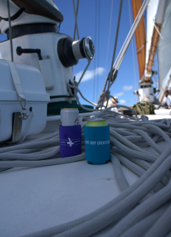 Lost Boy Koozie - Lost Boy Noun in Deep Teal — lifestyle shot, white Lost Boy Creations wordmark, on sailboat deck | Lost Boy Creations Key West