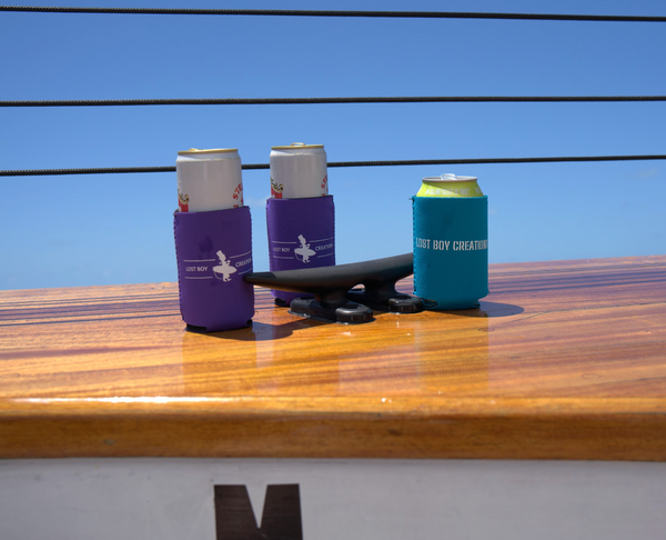 Lost Boy Koozie - Lost Girl Noun in Purple — lifestyle shot, white Lost Boy Creations surfer logo, on sailboat deck with teal Lost Boy Noun koozie | Lost Boy Creations Key West