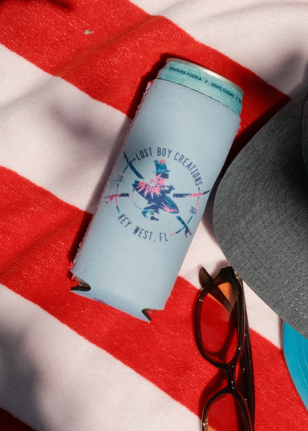 Lost Boy Koozie Floral in Light Blue — flat lay lifestyle on beach towel, multicolor floral Lost Boy Creations compass graphic in navy and pink | Lost Boy Creations Key West