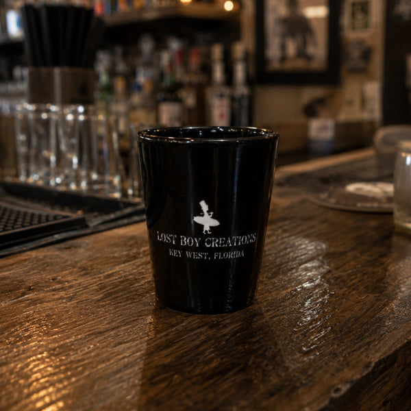 Lost Boy Creations LBC Shot Glass in black ceramic sitting on a wooden bar top showing Lost Boy Creations Key West Florida logo | Lost Boy Creations Key West