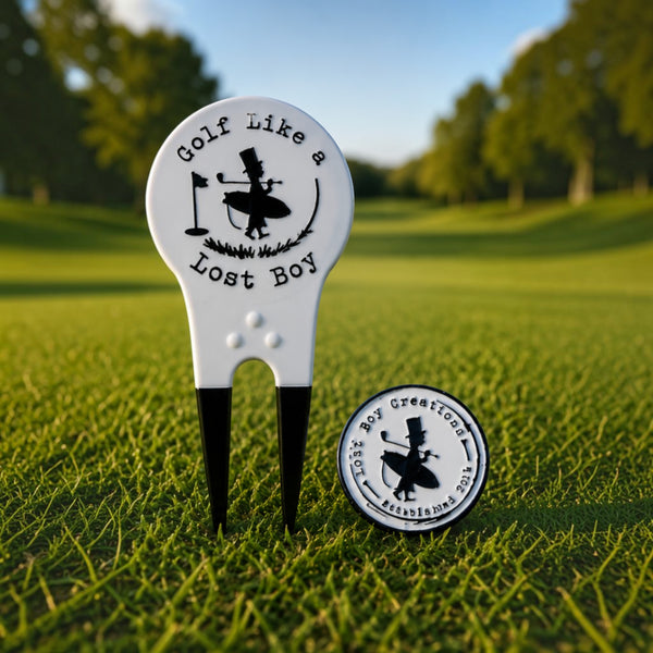 Lost Boy Creations white and black dual-prong divot tool and magnetic ball marker with golf graphic on green grass.