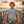 A young boy wearing a grey Lost Boy Creations Definition Tee on a wooden pier at sunset holding a surfboard and hat.