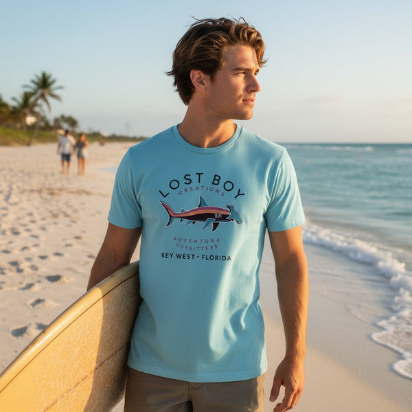 Man wearing Men's Hammerhead Tee light blue t-shirt with surfboard on sunny beach | Lost Boy Creations Key West