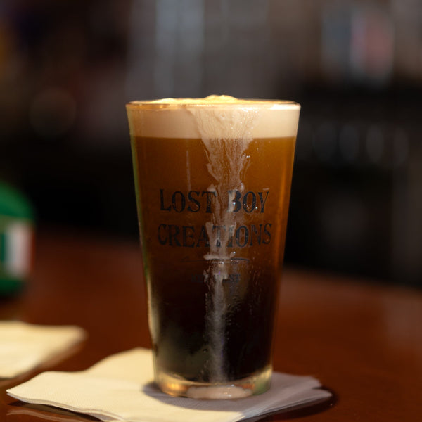 Lost Boy Creations Perfect Pint glass filled with dark beer and foam on a wooden bar top showing Lost Boy Creations logo | Lost Boy Creations Key West