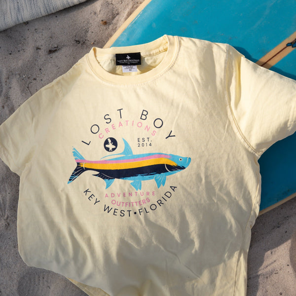 Lost Boy Creations Kid's Silver King Tee in lemon yellow laid on a surfboard showing colorful tarpon graphic and Adventure Outfitters Key West Florida text | Lost Boy Creations Key West